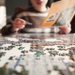 Boy sitting at a table working on a jigsaw puzzle while holding the paper.